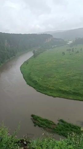The video shows a wide, meandering river flowing through a lush green valley. The riverbanks are lined with trees and vegetation, and in the distance, hills covered in forests rise up. The sky is overcast, suggesting a cloudy or rainy day. There are small scattered encampments or houses along the right bank of the river, and a few boats are visible on the water. The camera pans slowly across the landscape, offering a panoramic view of the natural scenery. The foreground shows a rocky, overgrown cliff with some wildflowers.