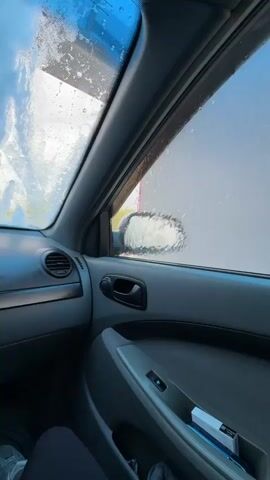 The video shows an interior view from a car, specifically from the passenger's seat, during a car wash in Khanskaya, Russia. The dashboard, passenger door panel with a visible door handle and window controls, and the passenger's feet in white sneakers are in view. A small white box, possibly a pack of cigarettes or gum, is tucked into the door pocket. Outside the car, a high-pressure water spray from a nozzle is directed first at the side window, then moves to the windshield, covering the glass with water and suds. A light-colored corrugated metal wall is visible outside the window before it becomes obscured. The time of day appears to be bright and sunny.