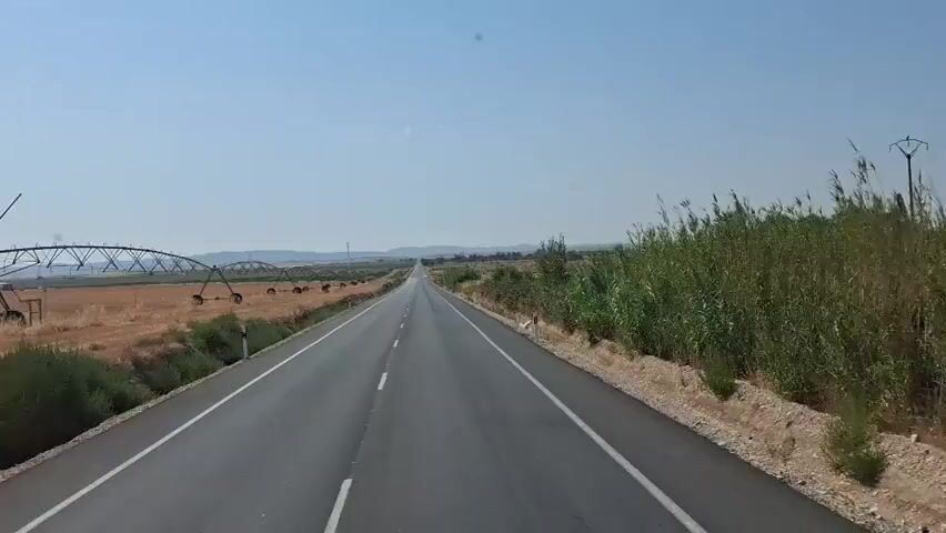 The video shows a highway stretching into the distance under a clear blue sky. The road is paved and has white lines marking the lanes. On the left side of the road, there are fields with agricultural irrigation equipment, resembling a large circular sprinkler system. Further in the distance, there are some trees and hills. On the right side of the road, there is dense vegetation, possibly reeds or tall grass, and then more dry, arid land with some scattered bushes and trees. Power lines and pylons are visible in the distance. The overall environment appears to be rural and agricultural, likely in a sunny climate given the clear sky and dry landscape. The video seems to be taken from a moving vehicle, providing a first-person perspective of the journey.