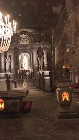 The video shows the interior of a church, possibly carved out of a salt mine, with a large chandelier dominating the left side of the frame. The walls are made of rough, dark brick or stone, speckled with what appears to be mineral deposits.  The church features a detailed altar area, housing what looks like religious statues. Candles are visible around the statue. The floor is tiled. Two people enter the frame, and one is taking pictures with a phone. The lighting is dim, with several light fixtures, including the chandelier, illuminating the space.