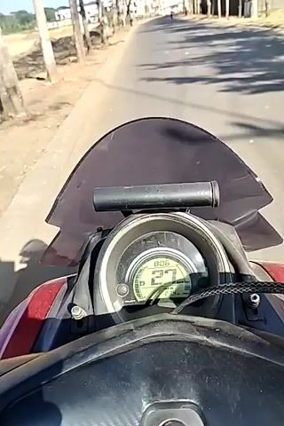 The video is filmed from the perspective of someone riding a motorcycle. The motorcycle's dashboard is visible in the foreground, showing a speedometer reading of 26. The road ahead is lined with trees on both sides. There are buildings and fields visible in the background. The sky is clear and blue. A scooter is visible on the road ahead. The motorcycle appears to be moving at a moderate speed. The lighting suggests it is daytime.