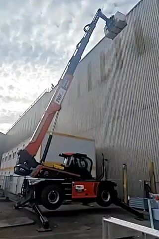The video begins by showing a close-up of a muddy, potholed concrete surface. The view then shifts to a telehandler, a type of construction equipment, positioned near a large, corrugated metal building. The telehandler's arm is extended upwards, lifting a white object. The scene appears to be an outdoor industrial or construction area.