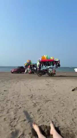 The video shows a sandy beach on a sunny day. In the foreground, there are two bare feet partially buried in the sand. The feet are pale and belong to someone lying down on the beach. In the background, there are two motorized tricycles parked near a red car. The tricycles are heavily decorated with colorful items, possibly balloons or other merchandise for sale. The ocean is visible in the distance, with a few people and boats scattered across the horizon. The sky is clear and blue.