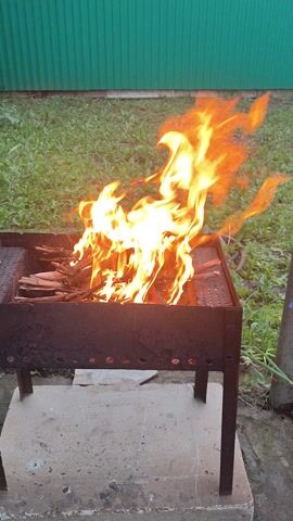 A time-lapse video shows a barbecue grill with wood catching fire. The grill is made of dark metal and sits on a concrete slab. To the left of the grill is a green corrugated metal fence, and behind it is a grassy area with some plants. The fire starts small and quickly grows, with flames reaching high into the air and flickering. The wood inside the grill appears to be burning vigorously. The video is shot from a slightly elevated angle, looking down at the grill. There are no people or animals visible in the video. The overall scene suggests an outdoor setting, possibly a backyard or patio.