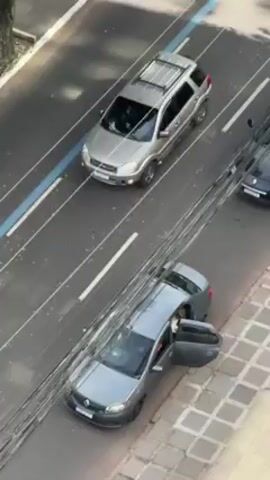 From an elevated perspective, the video captures a section of a multi-lane city street. A gray sedan is parked parallel to the sidewalk, its front and rear passenger doors open. At least one person is visible inside the car. Two vehicles, a silver SUV and a white compact car, are traveling in the lanes adjacent to the parked sedan. A third dark-colored car is partially visible in another lane. Utility wires are prominently seen in the foreground, obscuring the view of the street to some extent. The street has painted lane dividers including a blue line. The sidewalk is tiled with a geometric pattern.