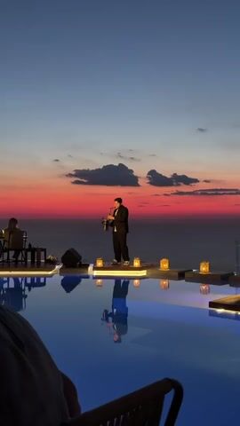 A man in a black suit and white t-shirt is playing a saxophone on a raised platform. He is standing in front of a body of water, possibly a pool, and a sunset is visible in the background. There are lanterns on the platform around him, and the water reflects the sky and the man. There are other people in the background, seated at tables. The overall atmosphere is elegant and serene.