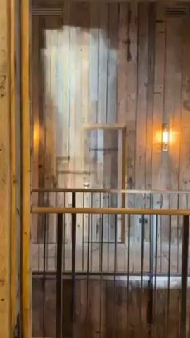 The video shows a view of a building interior with wood paneling on the walls.  A metal railing runs across the lower half of the frame, creating a sort of balcony or mezzanine.  There is a "CHANGING ROOM" sign on a door, centered between two light fixtures on the wall.  The light fixtures have a warm, yellowish glow. The overall impression is a rustic or industrial aesthetic.
