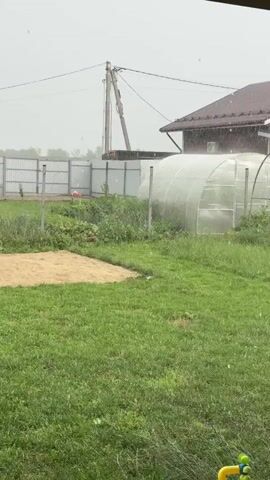The video shows a backyard scene during rainfall. A red-brown house with a white-trimmed window is visible in the background, along with a white greenhouse. A utility pole stands behind the house. In the foreground, green grass covers the yard, and a brown patch of soil is visible to the left. A dark-colored patio beam runs across the top of the frame, and a wooden support post is on the right. A red planter with a solar-powered garden light sits on the patio. Raindrops are visible falling through the air.