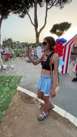 A young woman with brown hair and sunglasses stands on a paved area, holding a phone in her right hand and a bubble wand in her left. She is wearing a black crop top, a light blue denim skirt, and multi-colored sandals. The woman is smiling and looking at her phone as if recording a video or taking a picture. Bubbles are floating in the air around her and on a green grassy area to her left. In the background, several other people are visible, some walking and others standing. There are also trees and balloons in the background, suggesting an outdoor event or celebration. The lighting indicates it is daytime, likely late afternoon or early evening.