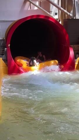 Two young girls in black swimsuits ride side-by-side in a double yellow inner tube. They emerge from a large red water slide into a pool of water. The girl on the left looks towards the camera, while the girl on the right looks at the girl on the left. The inner tube is partially submerged in the churning water. In the background, there is a yellow water slide structure and parts of a building interior with a ceiling.