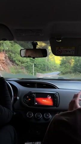 The video shows a first-person perspective from inside a car driving along a winding road through a densely forested area in Bratešići, Montenegro. The driver is visible on the left, with their hands on the steering wheel, simultaneously holding and interacting with a smartphone. The car's dashboard and infotainment system are in the foreground, with the screen displaying "Audio is off" in red text. A passenger's shoulder is partially visible on the right. Through the windshield, a narrow paved road can be seen disappearing into the distance, flanked by tall green trees and bright sunlight illuminating distant hillsides. The rearview mirror reflects a blurred image. A sun visor on the passenger side displays "AIRBAG" and a safety warning icon. The time of day appears to be sunny, likely late morning or afternoon.
