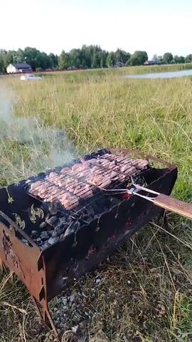 A close-up, low-angle shot shows food being grilled over coals in a metal barbecue pit. The pit is rustic, made of metal with decorative cutouts. A wire rack holds numerous pieces of raw meat, likely chicken, arranged in rows. Smoke rises from the grill, indicating it's actively cooking. In the background, a grassy field stretches out towards a body of water and a few buildings, suggesting an outdoor setting, possibly a park or rural area. The lighting is soft, indicating it may be late afternoon or early evening. There are no people visible in the frame, but a pair of tongs is resting on the grill.