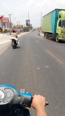 The video shows a first-person perspective of someone riding a blue motorcycle on a paved road. In front of the rider, another person is riding a motorcycle carrying a large white package on the back. In the background, there are buildings, trees, and utility poles. There are also other vehicles on the road, including a large green truck. The sky is visible and appears to be clear and sunny.