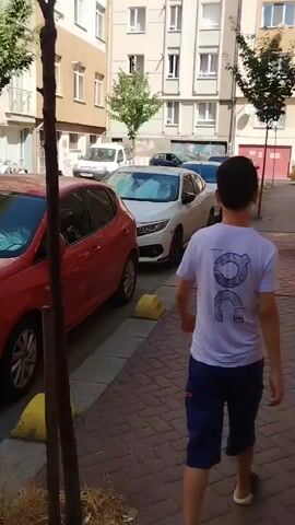 The video captures a street scene with parked cars and apartment buildings. A red car is prominently featured in the foreground, with a white car parked behind it. The street is lined with yellow parking bollards. In the background, several apartment buildings with balconies and windows are visible. Trees with green foliage are scattered throughout the scene, casting shadows on the ground. The sky is clear, suggesting a sunny day. The overall impression is of a typical urban environment on a bright day.