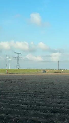 A light-colored car drives along a road that runs through a cultivated field. The field in the foreground appears to have been recently plowed, with dark, tilled soil forming parallel ridges. In the background, beyond the road and a strip of green grass, more cultivated fields are visible. Several wind turbines stand in the distance to the left of the frame, and a large electricity pylon is positioned closer to the road. The sky is blue with scattered white clouds. The shot seems to be taken from a moving vehicle, as evidenced by the car's side mirror visible in the bottom right corner of the frame.