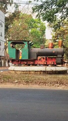 The video shows a static, weathered steam engine displayed outdoors on a circular concrete base. The engine is painted green and black, with red accents on its wheels and frame. Behind the engine is a backdrop of trees and foliage, suggesting a park or outdoor display area. To the left, a portion of a sign with graphic illustrations is visible. A red car drives past the engine from left to right in the frame during the video. The weather appears to be daytime with natural lighting.