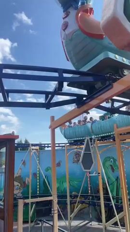 The video begins showing a close-up of a blue dinosaur-shaped roller coaster car against a cloudy blue sky. As the video progresses, the camera pans down and to the right, revealing the roller coaster track and other cars moving along it. Several people, appearing to be children and adults, are seated in the roller coaster cars. The roller coaster is operating at a fair or amusement park, evidenced by colorful signage and other rides visible in the background. The scene is outdoors on a sunny day.