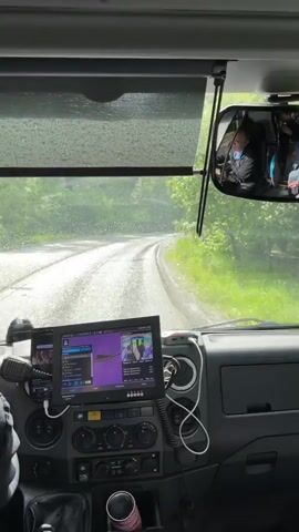 The video shows a wet winding road through a green wooded area as seen through the windshield of a vehicle. The windshield wipers are on and droplets of water are visible on the glass.  A monitoring screen on the dashboard shows a video of passengers inside the vehicle.  A small USB style device sits on top of the dashboard.  The road has worn asphalt showing its age. Other vehicles are visible on the road ahead.