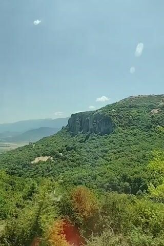 The video shows a panoramic view of a mountainous landscape under a clear blue sky with scattered clouds. In the foreground, there are lush green trees and dry, bare trees. In the mid-ground, a large, rugged rock formation stands out on a green, tree-covered mountainside. Further in the distance, a valley with a town or village can be seen, surrounded by rolling hills and mountains fading into a hazy blue. The camera appears to be moving, possibly from a vehicle, giving a sense of travel and perspective. The audio includes spoken words in a language that is not English, but the content is not discernible from the visual alone.