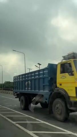 The video shows a view from inside a vehicle traveling on a road. The road has a barrier on the right side, made of concrete blocks with black and yellow stripes. There are streetlights along the road. Trees and vegetation are visible behind the barrier. The sky is overcast with dark clouds. In one frame, a blue truck with a cage-like structure on the back is seen driving on the road. The vehicle appears to be moving forward.