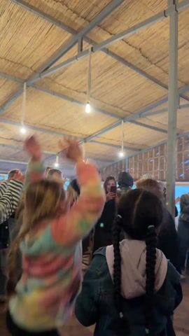 A group of people, including children, are gathered indoors under a light-colored, thatched roof. They are clapping, dancing, and some are taking pictures with their phones. Some of the adults are dressed in striped tops and vests. The space is illuminated by hanging light bulbs, and there are steel support beams. The overall atmosphere appears festive.