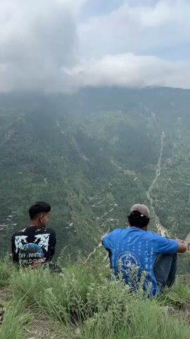 The video shows two individuals from behind, sitting on a grassy ledge overlooking a steep, verdant mountain valley. The person on the left wears a black t-shirt with white graphics, and the person on the right wears a blue t-shirt and a cap. Their legs are crossed or tucked beneath them as they face away from the camera and toward the expansive, cloud-covered mountain landscape. The valley floor is visible in the distance, with a winding road and some scattered buildings. The overall scene conveys a sense of peaceful contemplation amidst a natural, remote environment.