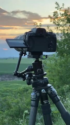 This video shows a DSLR camera mounted on a tripod in a natural setting, likely a viewpoint overlooking a canyon. The camera's LCD screen is tilted up, suggesting it is actively being used to frame a shot. The background depicts a vibrant sunset with colorful clouds painting the sky in hues of orange and pink. Below the sky, a winding canyon carved into green fields is visible, with steep rocky edges and lush vegetation. The overall scene conveys a sense of tranquility and the beauty of nature, captured through the lens of photography.