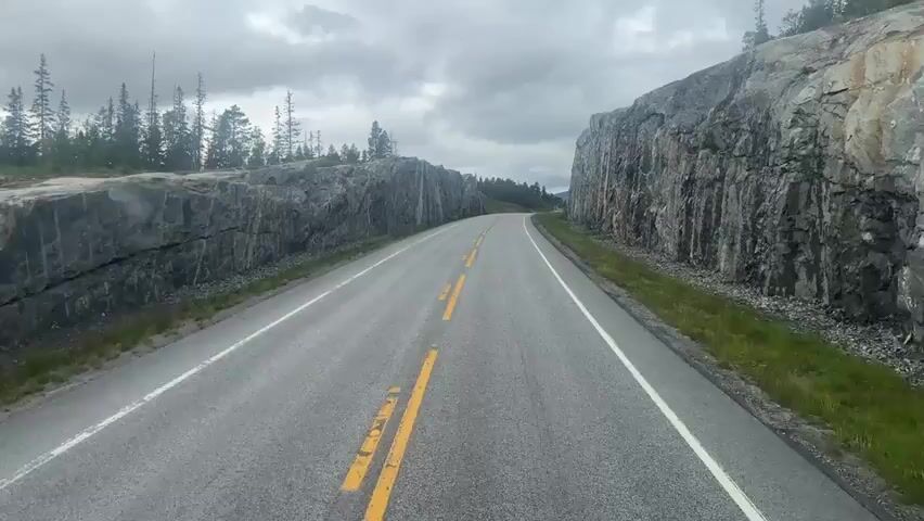 This is a first-person perspective video recording of a drive through a scenic mountain pass. The road is paved and marked with a double yellow line, indicating a two-way traffic lane. The driver is traveling uphill as the road curves to the right, leading into the distance.

Flanking the road on both sides are imposing, sheer rock faces. These cliffs are jagged and appear to be made of dark grey stone, with some lighter colored veins running through them. The textures of the rock are rough and weathered, suggesting a long geological history. On the left side of the road, sparse evergreen trees are visible growing on top of the rocky terrain. On the right side, the rocky slope gives way to a grassy verge that also appears somewhat sparse.

The sky above is overcast with heavy, grey clouds, hinting at potentially inclement weather or a generally cool climate. There are no visible people or vehicles in the immediate vicinity, contributing to a sense of solitude and emphasizing the vastness of the natural landscape. The lighting is diffused and consistent due to the cloud cover, suggesting it is daytime, likely mid-morning or afternoon. The overall atmosphere is one of natural grandeur and a quiet, solitary journey through a dramatic environment. No visible text is present in the video.