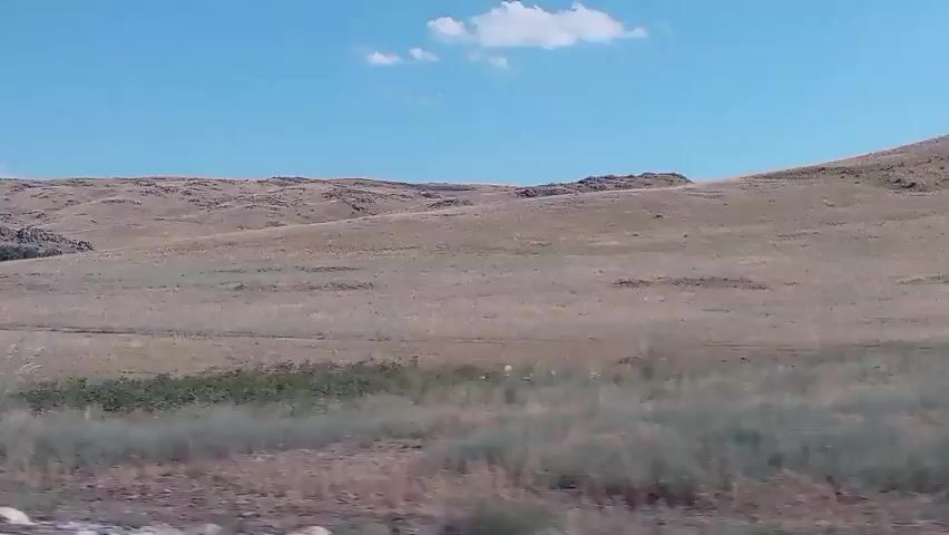 The video shows a wide, dry, hilly landscape under a clear blue sky with a few white clouds. The terrain is covered in sparse, dry grass and some scrubby bushes. There are rocky outcrops on the hills in the distance. The camera appears to be moving, possibly from a vehicle, as there is a slight blur and the foreground is slightly out of focus. A pole with a blue and white flag is visible on the right side of the frame. There are no people or animals visible in the scene. The overall impression is of a vast, arid, and undeveloped natural environment.