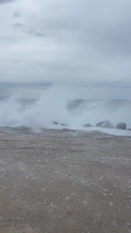 The video shows a choppy sea under a cloudy sky. Waves crash against rocks at the edge of a concrete shore.  Foam and spray from the waves reach the shore.  Mountains are visible in the distance on the horizon.