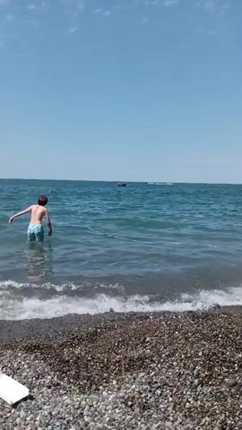 A young person with brown hair, wearing blue and white patterned swim trunks, stands in shallow water near a pebble beach. They are facing away from the camera towards the ocean. In the distance, a small boat or raft is visible on the water, with what appear to be other inflatable toys or watercraft further away. The sky is clear and blue, and the water is choppy.