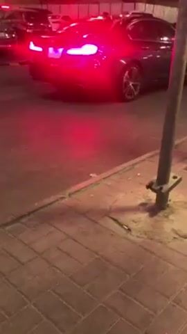 The video shows a street with parked cars on the left and in the distance. A dark-colored car with its rear lights illuminated is pulling away from the curb. There is a tall, gray pole visible on the right side of the frame. The ground is paved with square bricks. The scene is shot from a low angle, and the overall lighting suggests it is nighttime.