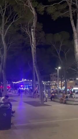 The video shows an outdoor scene at night with trees and artificial lighting. In the foreground, a person is sitting on a bench near a trash can, looking at their phone. Further back, a group of people are gathered, some sitting on benches and others standing. There is a stage with lights in the distance, suggesting an event or performance. A family with two children in a stroller is walking through the area, illuminated by green lights. The overall atmosphere appears to be lively, with people enjoying the evening.