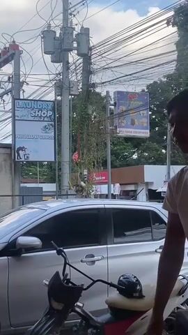 The video begins with a medium shot of a young man wearing a white t-shirt, looking to his left. The background shows a street scene with a silver car, a motorcycle, utility poles with numerous wires, and a laundry shop sign. As the video progresses, two women walk by, and a yellow tricycle passes in the background. The camera pans slightly to the right, revealing more of the street, including a food solutions sign and trees. The scene appears to be an outdoor urban environment with moderate traffic and pedestrian activity.