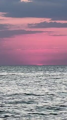A video shows a wide shot of a body of water, likely the ocean, with a sunset in the background. The sky is filled with clouds that are illuminated by the setting sun, creating vibrant shades of pink and purple. The sun itself is a small, bright line just above the horizon. The water is calm with gentle waves, and there is a line of buoys visible across the middle of the frame. The overall impression is one of a peaceful and beautiful natural scene.