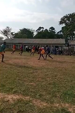 The video shows a group of young people playing a game on a grassy field in front of a building with a long, covered walkway. The players are wearing athletic attire, with some in yellow and orange tops and shorts, others in red and white, and one in a blue and green top. They appear to be playing a ball game, possibly soccer or a similar sport, as some players are running with the ball, kicking it, and one player in the center is jumping to hit the ball. There is a net or goal structure visible on the right side of the frame. Spectators are gathered on the left side, sitting and standing near the building. The weather appears to be overcast.
