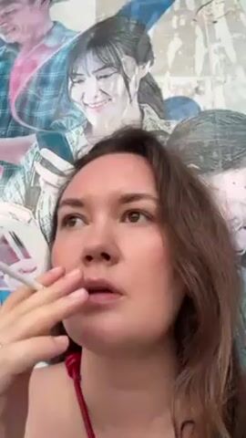 A woman with shoulder-length brown hair is smoking a cigarette. She is wearing a red top. In the background is a colorful mural of multiple people in a watercolor style. The woman is looking up and to the left. She has a ring on her finger. She takes a drag of her cigarette.