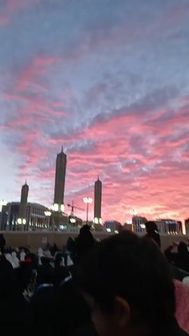 A vertical shot captures a crowd gathered outside the Masjid an-Nabawi mosque in Medina, Saudi Arabia. The camera focuses on a young girl in the foreground. She wears a grey headband and a black jacket. The sky above is a stunning blend of pink and white hues, painted across a sea of clouds. The mosque's minarets stand tall against this vibrant backdrop. In the midground, people dressed in dark clothing move about, some interacting with each other. The buildings surrounding the mosque are visible in the distance, and a construction crane is briefly seen near the center. The scene is peaceful, with the focus being on the beautiful sky and the religious site.
