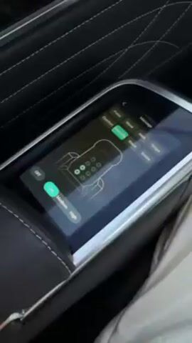 The video begins with a close-up shot of a car's armrest console, which features a touchscreen displaying massage options. The options include pulse, comfort, wave, walk, single, and shake. The screen also has low, medium, and high intensity settings. The camera then pans out to show the view from the car's window, revealing a snowy landscape with buildings and trees. The car appears to be moving. The environment outside includes snow-covered areas and overcast skies.