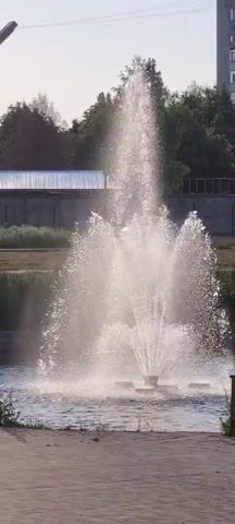 A fountain sprays water in a park. The fountain is located in a body of water, possibly a pond or lake. There are trees and buildings in the background. The sun is shining, casting a bright light on the scene. The water from the fountain is illuminated by the sun, creating a shimmering effect. The scene is peaceful and serene.