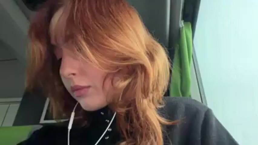 The video shows a person with wavy, ginger hair sitting and looking downwards. They wear a black top and white earphones. They use their left hand to move some hair from their forehead. The background suggests they might be on public transportation, with a window, a green curtain, and a digital display visible.