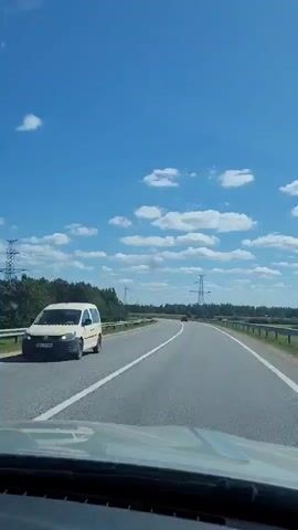 The video shows a view from inside a vehicle moving forward on a paved road. The road has white lines indicating lanes. On the left side of the road, there is a line of trees. On the right side, there is a flat, open landscape with more trees and power lines in the distance. The sky is blue with scattered white clouds. The sun appears to be shining. The vehicle appears to be the only one moving on this stretch of road, with a red vehicle visible in the far distance.