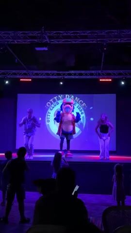 A costumed mascot, resembling a blue bird, stands center stage performing a dance routine. Two human dancers, a man on the left and a woman on the right, mirror the mascot's movements. They are on a raised stage illuminated by blue and red lights. Behind them, a large screen displays the "OTTY DANCE CLUB SINCE 1994" logo. In the foreground, an audience of silhouettes watches the performance. The environment appears to be an outdoor evening event, possibly a resort or entertainment venue.