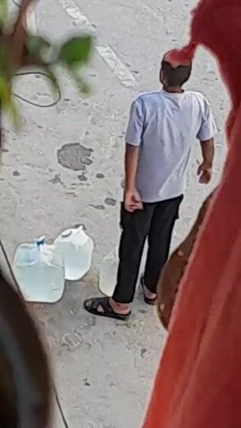 The video shows a young man standing next to three large plastic water jugs on a paved surface. He is wearing a light blue t-shirt, black pants, and black sandals. His arms are behind his back, and he appears to be looking down or to his left. The water jugs are partially filled with a clear liquid. The lighting suggests it is daytime. There is some debris or water stains on the ground around the jugs. In the foreground, out of focus, are parts of what appear to be people or fabrics.
