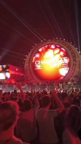 The video shows a large crowd of people at an outdoor music event, likely a concert or festival. In the background, a massive circular stage display shows the name "CHARLOTTE DE WITTE" in large white letters against a vibrant, fiery orange and red background with abstract shapes. The stage is illuminated by numerous bright white spotlights radiating from above. The crowd is densely packed, with many individuals raising their hands in the air, suggesting they are enjoying the performance. The lighting is predominantly red, casting a warm glow over the audience and the surrounding structures. The overall atmosphere appears energetic and festive.