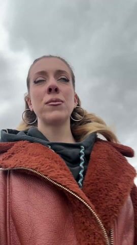 Here is a brief description of the video:
A woman wearing a rust-colored coat, black hoodie, and large hoop earrings is walking through a parking garage with a cloudy sky overhead. She looks into the camera and makes a few different faces. Cars are visible in the background.