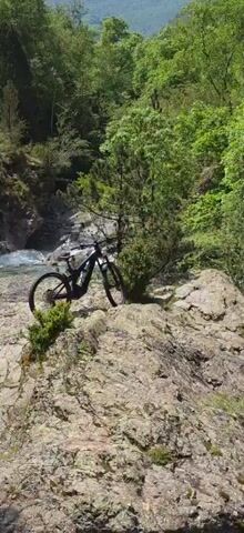 The video shows a scene on a rocky outcrop overlooking a fast-flowing river. At the beginning of the video, a mountain bike is parked on the rock. The camera pans down to reveal the river below. The water is a turquoise color and is rushing through a narrow gorge. Trees line the banks of the river.