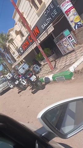 The video shows two motorcycles parked on a sidewalk in front of a cafe. The cafe is called "O Bon Coin Salon De". One motorcycle is silver with several silver storage boxes attached to the back, and the other is white, blue and red with a large windshield. The video is taken from the perspective of someone in a car, as a car door and side mirror are visible.  The sidewalk is painted with alternating green and white stripes. The pavement appears to be made of sandy gravel. The day is bright and sunny.