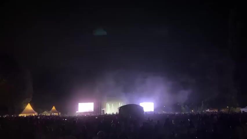 The video shows a crowded outdoor concert at night. A stage with bright lights and smoke is visible in the distance. Large screens flank the stage, displaying abstract pink and purple visuals. The audience is densely packed, with many people holding up their phones. Tents with yellow roofs are set up on the left side of the frame. Trees form a dark silhouette behind the stage. The overall atmosphere is dark with intermittent bursts of light from the stage.