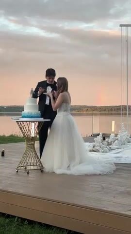 A bride in a white wedding dress and a groom in a black tuxedo cut a three-tiered white and blue cake. The cake has small figurines on top. The couple stands on a wooden deck next to a small gold table holding the cake. A body of water is visible behind them, with trees on the opposite shore. A faint rainbow is visible in the sky. The sky is filled with clouds tinted with sunset colors. To the right, a white decorated archway and a white fabric covering the ground suggest a wedding ceremony or reception setup.